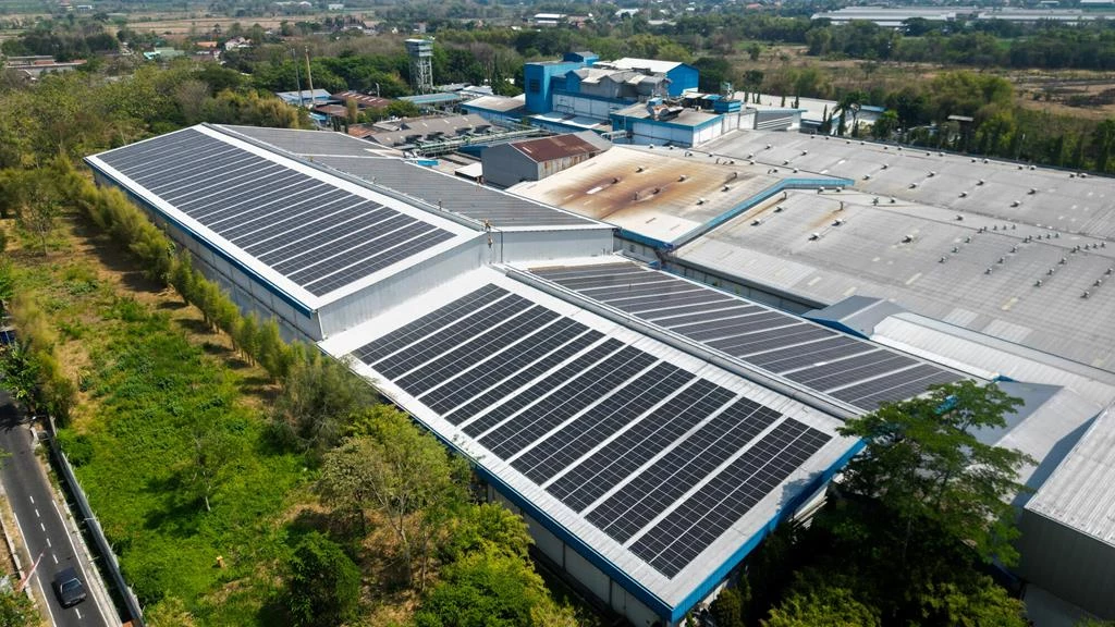Vista aerea di capannoni industriali con tetti bianchi coperti da pannelli solari, tra alberi e una strada asfaltata